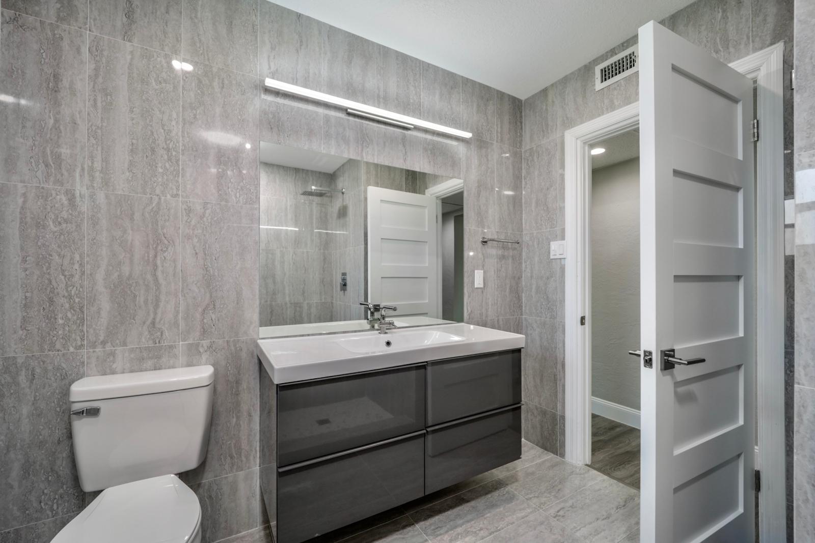 Travertine-tile bathroom with floating dark vanity and large mirror — The Travertine Suite, Palm Coast, FL